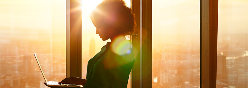 Decorative image showing a Black woman standing in front of large windows in an office building, silhouetted by sunlight as she gazes at the screen of a laptop.